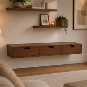 May include: A long, rectangular, wall-mounted wooden console with three drawers, positioned below two floating shelves. The top shelf holds a potted plant and books, while the lower shelf features a framed picture and a small candle.