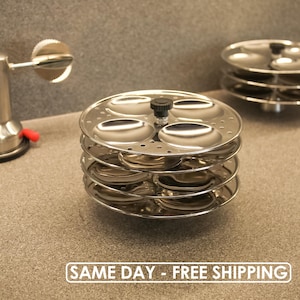 Könnte beinhalten: Ein Stapel glänzender Idli-Dampfplatten aus Edelstahl mit einem zentralen schwarzen Knopf. Die Platten haben mehrere kreisförmige Vertiefungen. Der Text "SAME DAY - FREE SHIPPING" befindet sich am unteren Rand des Bildes. Ein weiterer Idli-Dampfgarer befindet sich im Hintergrund.