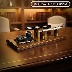 May include: A wooden desk set featuring a black inkwell, two pens, and pen holders. The pens are black and silver, and the inkwell is square. The set is displayed on a wooden tray with a dark finish.
