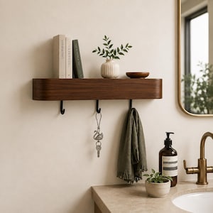 Könnte beinhalten: Ein schwebendes Regal aus dunklem Holz mit drei schwarzen Haken, das Bücher, eine kleine weiße Vase mit Grün und eine kleine Holzschale enthält. Ein grünes Handtuch und ein Schlüsselanhänger hängen darunter. Das Regal ist an einer hellen Wand befestigt.