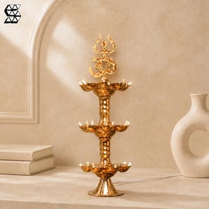 Op de afbeelding: Een goudkleurige metalen etagère-lamp met drie niveaus van kleine, komvormige lichthouders. De bovenkant van de lamp is versierd met een decoratief Om-symbool en een drietandvormige top. De structuur rust op een ronde voet.