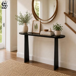 May include: A black console table with a long, narrow, oval-shaped top and two wide, tapered legs. The table holds a beige ceramic vase with green foliage, a framed fern print, a stack of three books, a small wooden bowl, and a potted plant.