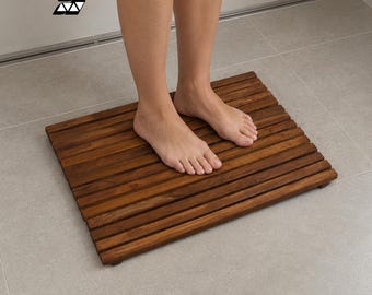 Op de afbeelding: Een rechthoekige bruine houten badmat bestaande uit parallelle latten die horizontaal zijn gerangschikt. De mat heeft een donkere houtnerfafwerking en ligt op een lichtgrijze tegelvloer. Een logo met een gestileerde letter S is zichtbaar.