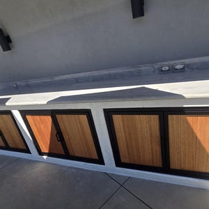 May include: Outdoor kitchen island with a concrete worktop and three wooden cabinet doors. The doors have black frames and handles. The island is set against a light grey wall.