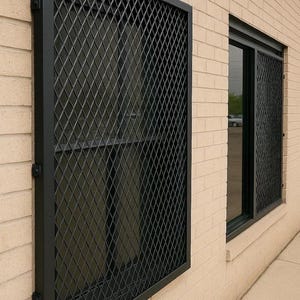 Puede incluir: Dos ventanas con pantallas de seguridad negras con diseño de diamantes. Las pantallas están montadas en una pared de ladrillo beige claro. Una ventana es más grande y tiene una pantalla completa, la otra es más pequeña con una pantalla parcial.