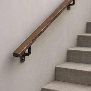 Puede incluir: Una barandilla de color bronce está montada en una pared gris claro, con soportes de metal negro. La barandilla está colocada en diagonal, sobre una escalera de hormigón gris. El diseño es moderno y minimalista.