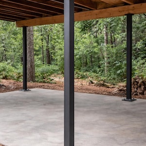 May include: Exterior view of a covered patio with a concrete floor and dark gray metal support columns. The ceiling is made of wooden planks. The background features a lush green forest.