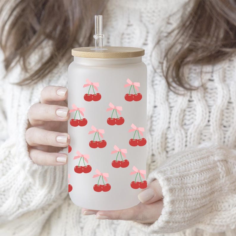 May include: A frosted glass water bottle with a bamboo lid and a straw. The bottle has a repeating pattern of red cherries with pink bows.