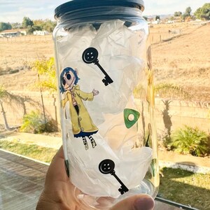 以下が含まれることがあります： 黒い蓋が付いた透明なガラス瓶。白いティッシュペーパーが詰められており、カラフルなステッカーで飾られています。ステッカーには、青い髪と黄色のコートを着た漫画のキャラクター、鍵、ボタンが描かれています。