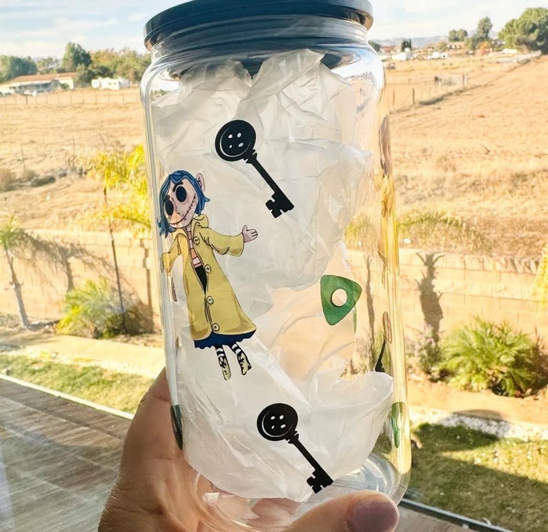 以下が含まれることがあります： 黒い蓋が付いた透明なガラス瓶。白いティッシュペーパーが詰められており、カラフルなステッカーで飾られています。ステッカーには、青い髪と黄色のコートを着た漫画のキャラクター、鍵、ボタンが描かれています。