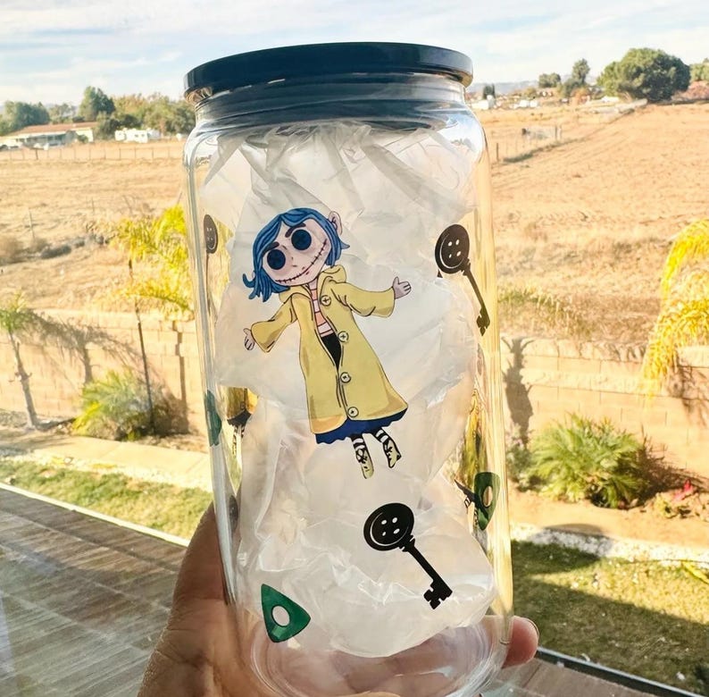 以下が含まれることがあります： 黒い蓋が付いた透明なガラス瓶に、青い髪と黄色のレインコートを着た漫画のキャラクターが描かれています。キャラクターは、鍵、ボタン、三角形など、さまざまなステッカーで囲まれています。