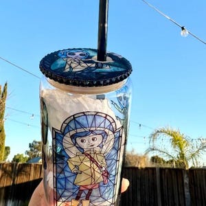 Puede incluir: Un vaso de vidrio transparente con una pajita negra y una tapa con un dise&ntilde;o de personaje de dibujos animados. El vaso tiene una ilustraci&oacute;n de estilo vidriera de un personaje con un abrigo amarillo. La tapa y el dise&ntilde;o del vaso tienen una combinaci&oacute;n de colores azul y negro.