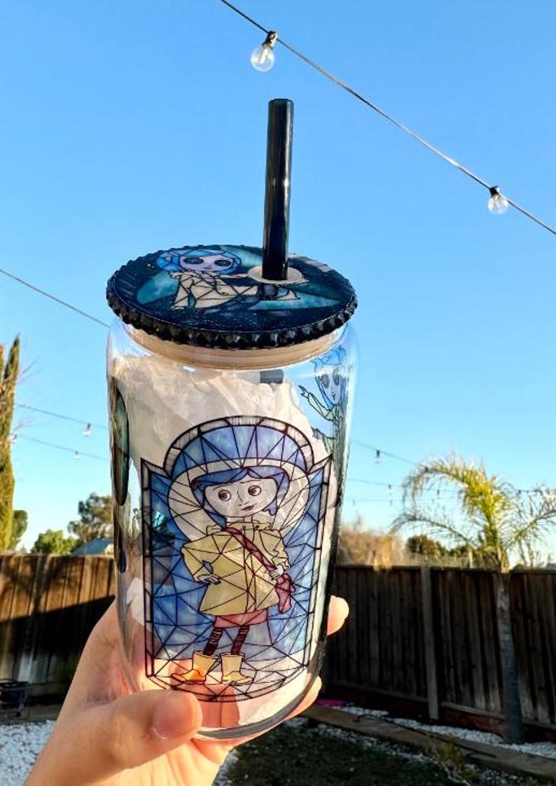 Puede incluir: Un vaso de vidrio transparente con una pajita negra y una tapa con un dise&ntilde;o de personaje de dibujos animados. El vaso tiene una ilustraci&oacute;n de estilo vidriera de un personaje con un abrigo amarillo. La tapa y el dise&ntilde;o del vaso tienen una combinaci&oacute;n de colores azul y negro.