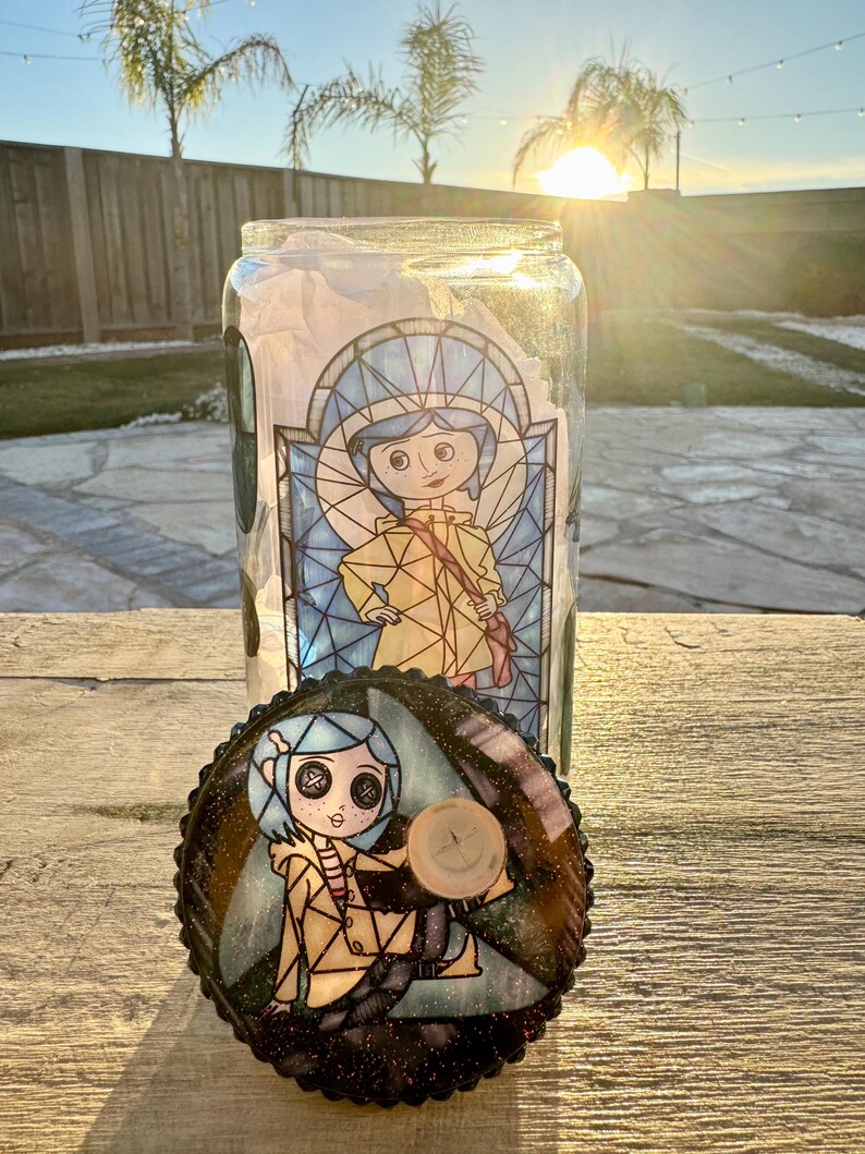 Puede incluir: Un tarro de cristal transparente con una tapa decorativa con un dise&ntilde;o de personaje de dibujos animados. El tarro tiene una ilustraci&oacute;n de estilo vidriera de una ni&ntilde;a con un abrigo amarillo. La tapa tiene un dise&ntilde;o a juego con un bot&oacute;n de madera.