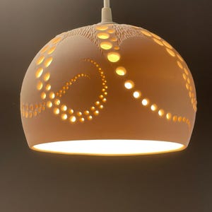 May include: A beige spherical pendant light. The light features a pattern of circular cutouts, allowing light to pass through. It hangs from a white cord. The light's diameter is approximately 25 cm.
