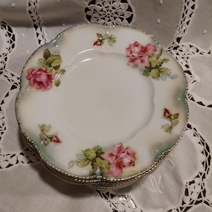 May include: A stack of white porcelain plates with scalloped edges and a gold-trimmed rim. The plates feature a floral pattern of pink roses, green leaves, and small rosebuds scattered around the border, with a soft green wash on the edges.