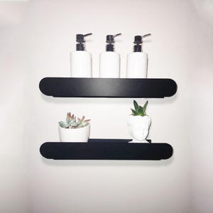 May include: Two black floating shelves with rounded edges, mounted on a white wall. The top shelf has three white pump bottles with black tops. The bottom shelf has a white planter with a succulent plant and a white planter shaped like a human head with a succulent plant.