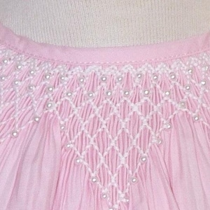 May include: Close-up of a light pink garment with intricate white smocking and delicate pearl-like beads. The smocking forms a diamond pattern across the chest and shoulders, adding texture and detail to the soft fabric. The garment has a round neckline.
