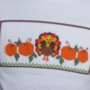 May include: A white shirt featuring a rectangular smocked panel with brown trim. The panel displays an embroidered turkey in the center, flanked by two orange pumpkins on each side, all accented with green leaves and a green zigzag border.