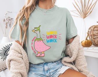 May include: A light green t-shirt featuring a cartoon duck dressed as a strawberry. The duck has a green hat and pink boots. The shirt has the words "WORK WORK WORK" in yellow, pink, and blue, and "TOO BUSY TO QUACK" in black.
