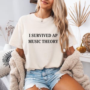 May include: A cream-coloured t-shirt featuring the black text "I SURVIVED AP MUSIC THEORY". The shirt is a relaxed fit, suitable for casual wear. The image shows a person wearing the t-shirt.