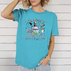 May include: A turquoise t-shirt featuring a colorful graphic design. The design includes a tequila bottle, margarita glass, and the words "Midnight Margaritas". The shirt is a short-sleeved crew neck.