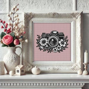 May include: A framed cross-stitch art piece featuring a black camera design surrounded by white flowers on a pink background. The frame is ornate and white. The image is displayed on a white mantel with decorative items and a vase of flowers.