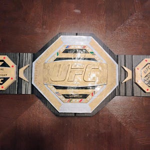 Op de afbeelding: Een gouden en zwarte UFC World Championship-riem met de tekst "UFC" en "Wereldkampioen" erop. De riem is versierd met een zwart-wit patroon en heeft een zilveren achthoek in het midden.