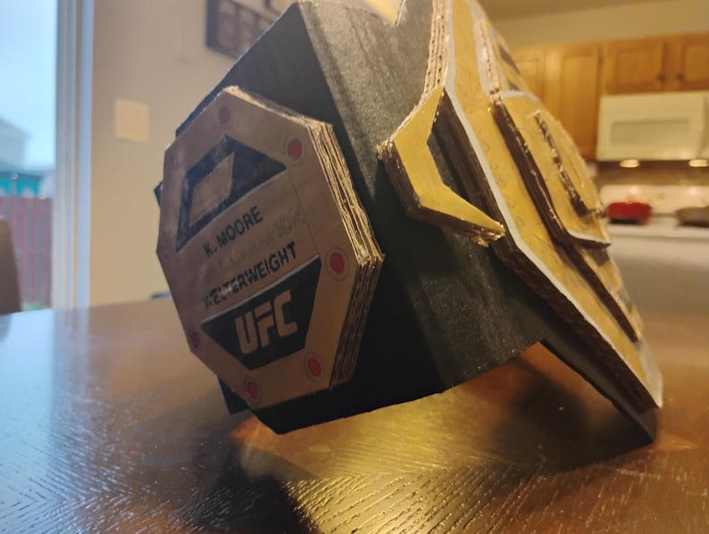 Op de afbeelding: Een kartonnen replica van een UFC-kampioensgordel met een goud-zilveren ontwerp. De gordel heeft de tekst "K. MOORE" en "UFC" samen met de woorden "Weltergewicht Kampioen".