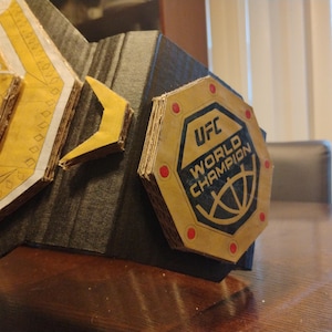 Op de afbeelding: Een kartonnen replica van de UFC World Champion-riem. De riem is goudkleurig met zwarte en rode accenten. De middelste plaat heeft het UFC-logo en de woorden "World Champion".