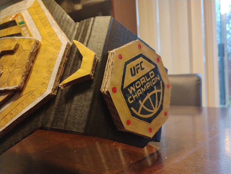 Op de afbeelding: Een kartonnen replica van de UFC World Champion-riem. De riem is goudkleurig met zwarte en rode accenten. De middelste plaat heeft het UFC-logo en de woorden "World Champion".