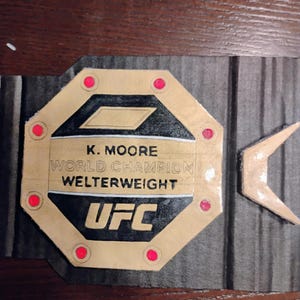 Op de afbeelding: Een gouden UFC-kampioensgordel met rode accenten. De gordel heeft de tekst "K. Moore Wereldkampioen Weltergewicht UFC".