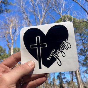 May include: A white square sticker featuring a black heart with a cross and the word "Jesus" in a cursive font. The sticker is held up against a backdrop of trees and a blue sky.