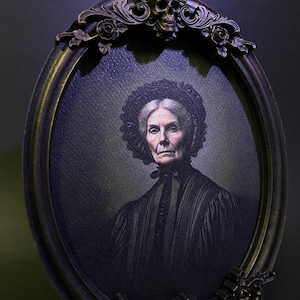 May include: An oval portrait in a black ornate frame with a skull at the top. The portrait is of an older woman with grey hair wearing a black dress and a lace cap.