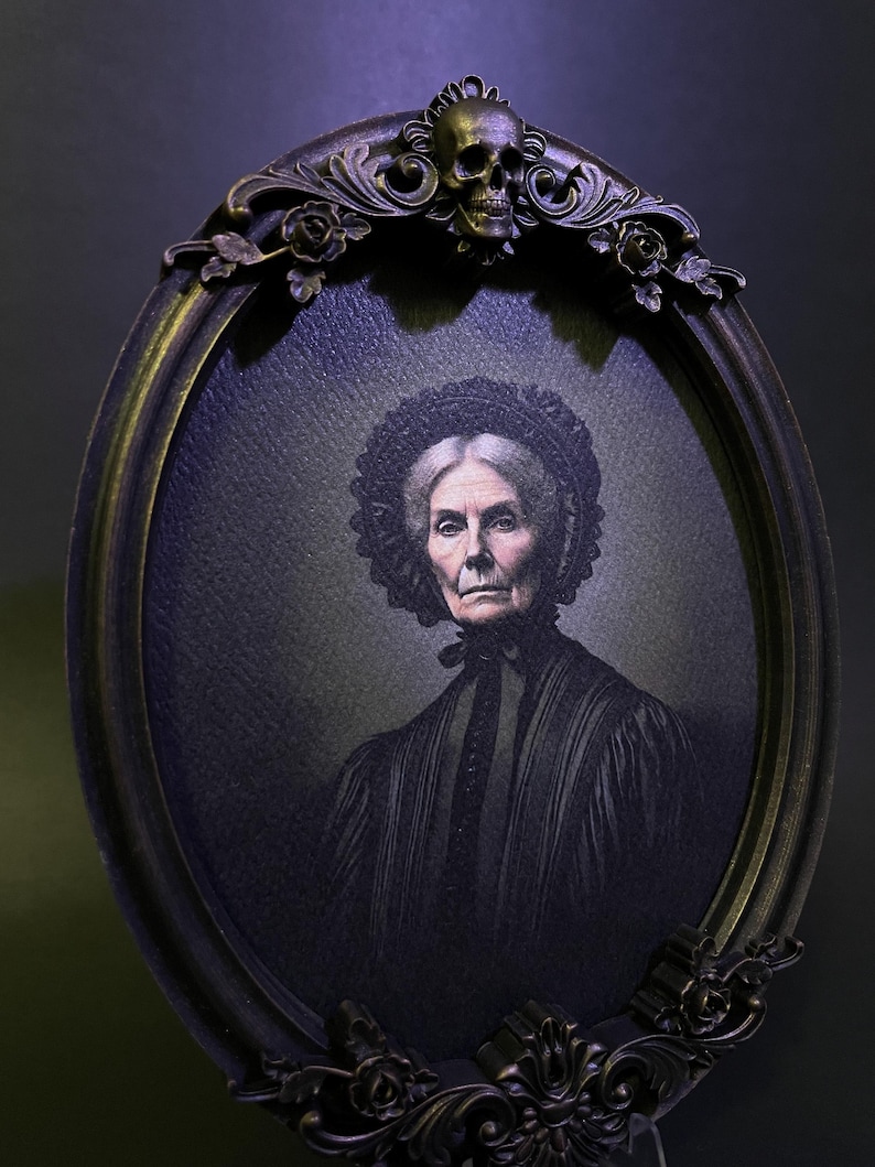 May include: An oval portrait in a black ornate frame with a skull at the top. The portrait is of an older woman with grey hair wearing a black dress and a lace cap.