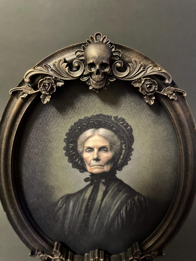 May include: An oval portrait of an elderly woman in a black dress and a lace bonnet, framed in a dark brown ornate frame with a skull and floral accents.
