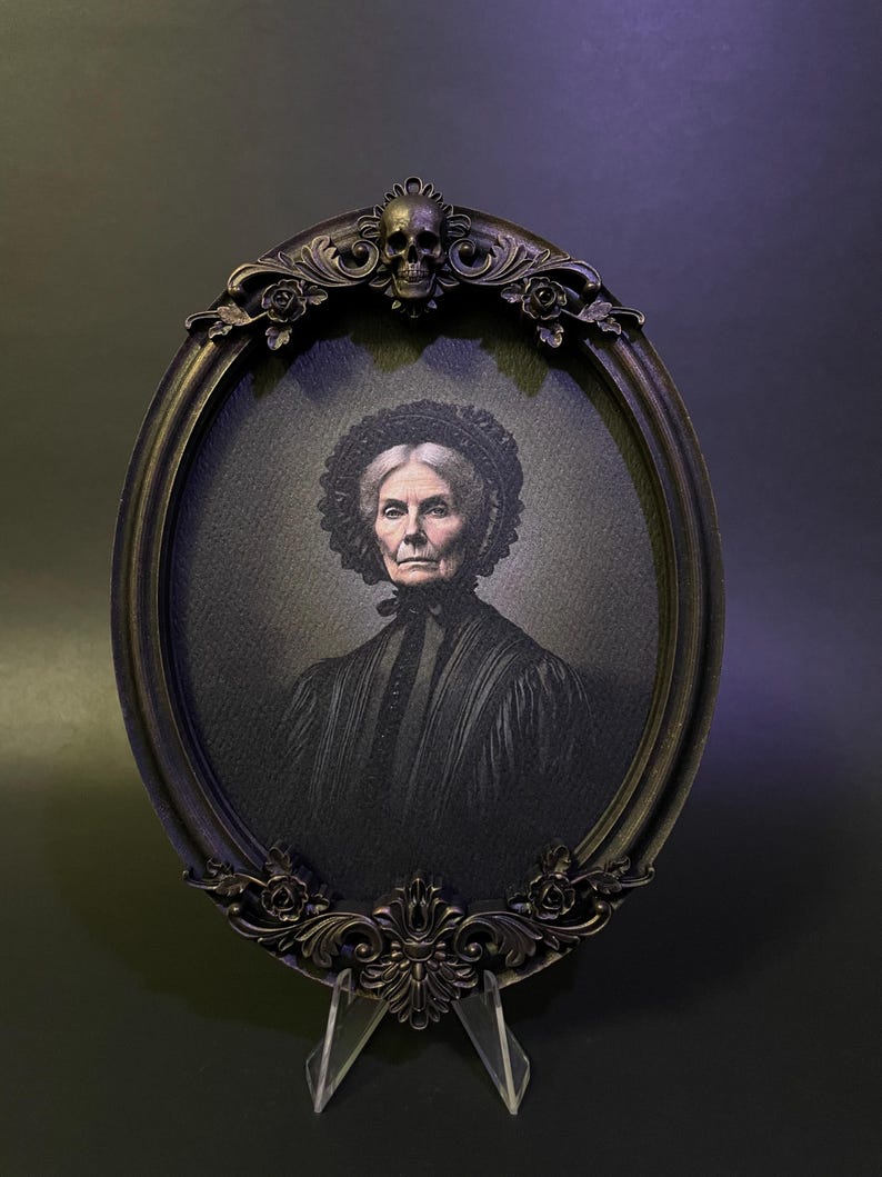 May include: An oval portrait of an older woman in a black dress with a lace collar, framed in a black ornate frame with a skull at the top.