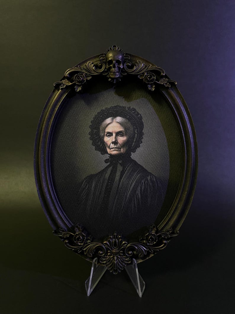 May include: A black oval frame with ornate details, including a skull at the top, holds a black and white portrait of an older woman wearing a black dress and a lace head covering.
