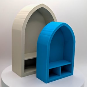 May include: Two decorative arched shelves, one in a light beige color and the other in bright blue. Each shelf has a rectangular base with a shelf inside. The shelves are displayed on a white surface.