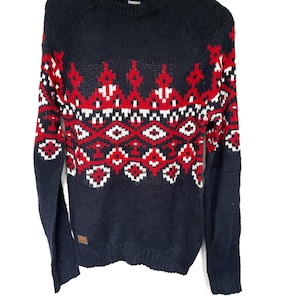May include: A navy blue knitted jumper featuring a red and white geometric design across the chest and sleeves. The jumper has a crew neck and long sleeves. A small brown label is sewn on the lower left side.