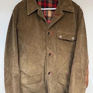 May include: A brown corduroy jacket featuring a pointed collar, a button-front closure with leather-reinforced accents, two lower pockets, and a single chest pocket. The interior is lined with a red, green, and tan plaid fabric pattern.