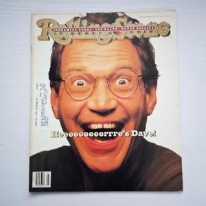 Puede incluir: Portada de la revista Rolling Stone con un retrato de un hombre con gafas, sonriendo ampliamente. El titular dice "Rolling Stone" en fuente dorada. Debajo de la imagen está el texto "Heeeeeeeeerrrre's Dave!"