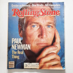 Puede incluir: Una portada de la revista Rolling Stone vintage con Paul Newman. La portada es azul y blanca con el nombre de la revista en rojo y negro. El titular dice "Paul Newman The Real Thing".