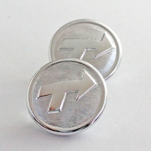 Puede incluir: Dos botones plateados brillantes con un diseño de flecha en relieve que apunta hacia la derecha. Los botones redondos tienen un borde texturizado y están sobre un fondo blanco. Estos botones podrían usarse para ropa o manualidades.