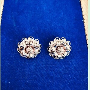 May include: A pair of silver-toned flower-shaped stud earrings. Each earring features a detailed floral design with multiple petals and a textured center. The earrings are displayed on a blue surface.