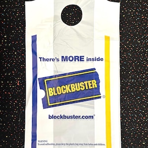 May include: White plastic Blockbuster bag with a handle and a circular hole. The bag features the Blockbuster logo in blue and yellow, with the text "There's MORE inside" and "blockbuster.com". A warning is printed at the bottom.