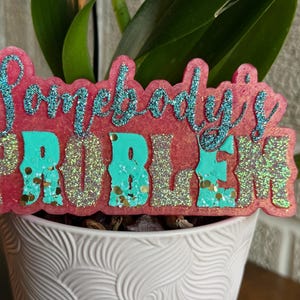 May include: A decorative sign featuring the text Somebody's PROBLEM in a layered design. The word Somebody's is in blue glitter script, while the word PROBLEM is in large, blocky, teal and silver glitter letters on a pink textured base.