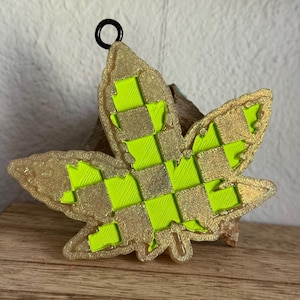 May include: A green and gold checkered cannabis leaf ornament with a black metal loop for hanging.