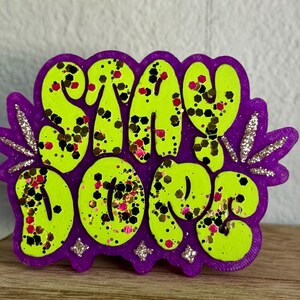 May include: A purple and lime green pin with the words "STAY DOPE" in bubble letters. The letters are lime green with black, red, and gold glitter. The pin has a purple border with silver glitter accents.