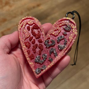 May include: A heart-shaped ornament with a glittery pink surface and a black string for hanging. The ornament features a leopard print design in red and silver glitter. The edges are also outlined with glitter.